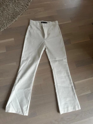 Vita kostymbyxor från Zara - Säljer ett par stilrena vita kostymbyxor från Zara. Byxorna har raka ben med lätt utsvängning, hög midja och fejkade fickor fram. Materialet känns mjukt och har en lätt stretch för extra komfort. Perfekta för en clean och trendig look.