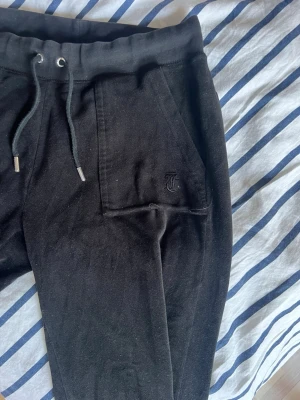 Juicy Couture  - Svarta mjukisbyxor från Juicy Couture med broderad logga på fickan. Byxorna har dragsko i midjan, två sidofickor fram och två bakfickor med knapp. Tillverkade i mjukt material för maximal komfort och avslappnad passform.   Två små hål i fickorna där fram, går enkelt att sy. Det små knapparna där bak, lite slitna.