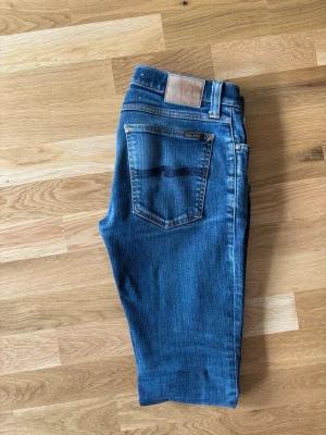 Blå jeans från Nudie Jeans - Säljer ett par klassiska blå jeans från Nudie Jeans i slimmad passform. Storlek 30/32. Jag på bilden är 180 lång. Börjar slitas lite mellan benen som man ser på bilden, men inga hål.