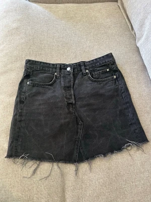 Svart jeanskjol med rå kant - Cool svart jeanskjol med rå, fransig kant nertill och klassiska fem fickor. Kjolen har knapp- och dragkedjestängning framtill och är i en rak, kort modell. Perfekt för en avslappnad och trendig look.