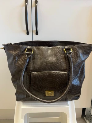 Mörkbrun Don Donna väska, ormmönstrat skinn - Snygg mörkbrun handväska i ormmönstrat konstläder med två rejäla handtag och guldfärgade metalldetaljer. Har tecken på användning t.ex insidan är nopprig och få lösa trådar men annars fullt fungerande. Ca 28cm hög och 41cm bred.