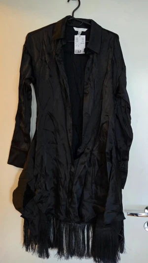 Svart långklänning med fransar H&M med prislapp - Svart långklänning från H&M i glansigt material med långa ärmar och knappar framtill. Klänningen har en asymmetrisk nederkant med långa fransar som ger en unik look samt möjlighet att knyta framtill. Perfekt för dig som vill sticka ut med en cool och edgy stil.