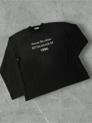 Svart långärmad topp Acne Studios | M  - Svart långärmad topp från Acne Studios med vit text 'Acne Studios STOCKHOLM 1996' på bröstet. Klassisk rund halsringning och rak passform. Perfekt för en stilren och avslappnad look.