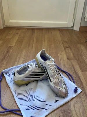 Adidas F50 - Säljer ett par Adidas F50 fotbollsskor i vitt med svarta och lila detaljer. Skorna har snörning, låg profil och FG-dobbar för spel på naturgräs. Ovandelen är i syntetmaterial och sulan har en gul ton. Kommer med tillhörande dustbag. Pris kan diskuteras