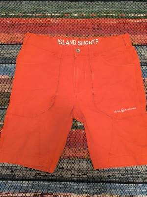 Orange shorts från Sail Racing - Snygga shorts från Sail Racing i modellen "Island Shorts". Klassisk seglardesign med rak passform och längre ben. Super praktiska med stora fickor. Färgen är en distinkt orange-röd ("spice"/korall-ton).  Skick: Använt men gott skick. Lite snyggt solblekta. Notera liten fläck på framsidan (se bild) – i övrigt fräscha.