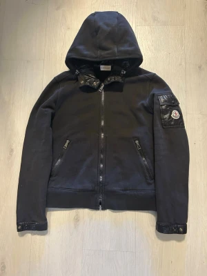  Moncler cardigan - Snygg svart hoodie från Moncler med dragkedja framtill, två fickor med dragkedja och en ikonisk logotyp på ärmen. Huva och detaljer i nylon på ärmen ger en exklusiv känsla. Perfekt för dig som gillar streetwear och premiumkänsla.    Womens size M fits S-M