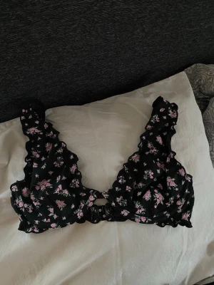 Svart blommig bikini topp med byglar under bysten.  - Säljer en svart bikini topp med rosa och vita blommor och volangdetaljer på axelbanden. Bh:n har breda axelband och knäppning fram, samt justerbara band i ryggen. Materialet känns mjukt och stretchigt, perfekt för en solig sommar dag. 