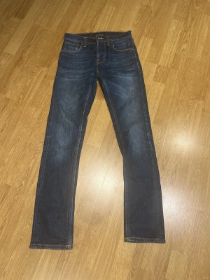  Nudie Jeans grim trim - Snygga mörkblå jeans från Nudie Jeans med klassisk femficksdesign och raka ben. Jeansen har kontrastsömmar och subtila slitningar för en cool look. Tillverkade i denim med en autentisk känsla och detaljerade bakfickor med Nudie-logga. Storlek 28/32