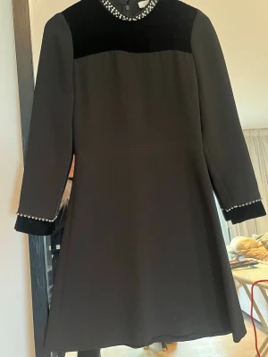 Sandro Paris klänning  - Elegant svart klänning med lång ärm och strass detaljer vid halsringning och ärmslut. Klänningen har en klassisk A-linjeform och sammetsliknande partier upptill och ärmslut.