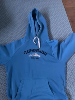 Blå Peak Performance hoodie - Snygg blå hoodie från Peak Performance med tryck framtill och vit dragsko i huvan. Klassisk känguruficka och mjukt material, perfekt för en avslappnad stil. Peak Performance-logga och bergmotiv på bröstet. Passar dig som gillar sportig och casual look. Priset går väl att diskuteras 