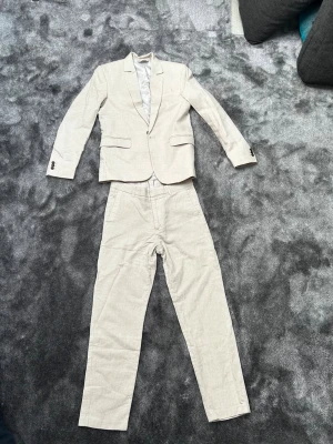 Beige kostym i linne - Snygg beige kostym med kavaj och byxor i linne från H&M. Kavajen har klassisk passform, två knappar och fickor med lock. Perfekt för dig som vill ha en stilren och fräsch look. Byxorna har raka ben och enkel design.