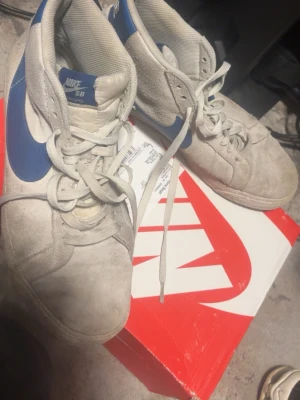 Nike SB Blazer Mid vita/blå sneakers - Nike SB Blazer Mid sneakers i vitt mockaläder med blå swoosh och detaljer. Klassisk siluett med högt skaft, vit yttersula och gummisula med fiskbensmönster. Perfekta för dig som gillar streetstyle och skate.