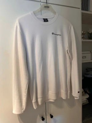 Vit sweatshirt från Champion - Vit sweatshirt från Champion med broderad logga på bröstet och liten logga på ärmen. Klassisk rund halsringning och ribbade muddar vid ärmslut och nederkant. Perfekt för en clean och sportig look.