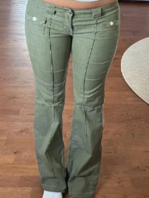 Gröna bootcut jeans med fickor - Snygga gröna bootcut jeans med markerade sömmar framtill och två fickor med lock och knapp. Jeansen har hällor för skärp och stängs med knapp och dragkedja. Perfekta för en trendig och avslappnad stil.