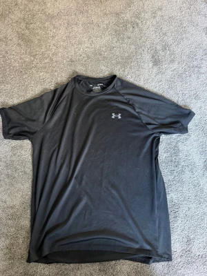 Svart t-shirt från Under Armour - Svart t-shirt från Under Armour med diskret logga på bröstet. Tillverkad i ett lätt och funktionellt syntetmaterial som andas, perfekt för aktiva dagar. Klassisk passform med korta ärmar och rund hals.