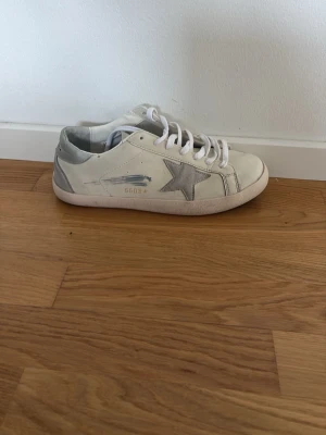 Vita sneakers från Golden Goose -  vita sneakers från Golden Goose med grå stjärndetalj på sidan och slitna effekter. Skorna har vita skosnören. Tillverkade i läder med en klassisk rund tå och grå detaljer på hälen. 