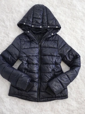 Vintage jacka från zalando - S - Snygg svart pufferjacka från Defacto med quiltad design och dragkedja framtill. Jackan har en stor huva med tryckknappar och ribbade muddar vid ärmsluten. Perfekt för kalla vinterdagar och riktigt skön att bära. Materialet är glansigt och vattenavvisande.