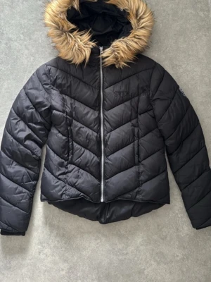 Svart pufferjacka med fuskpäls från SVEA - Snygg svart pufferjacka från SVEA med broderad logga på bröstet. Jackan har dragkedja framtill, huva med beige fuskpäls och ett quiltat mönster i V-form. Perfekt för kalla dagar och riktigt varm.