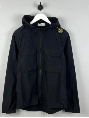 Svart vindjacka från Stone Island - Snygg svart vindjacka från Stone Island med ikonisk patch på ärmen. Jackan har huva, två stora bröstfickor med lock och dragkedja framtill. Tillverkad i lätt och slitstarkt material, perfekt för blåsiga dagar.