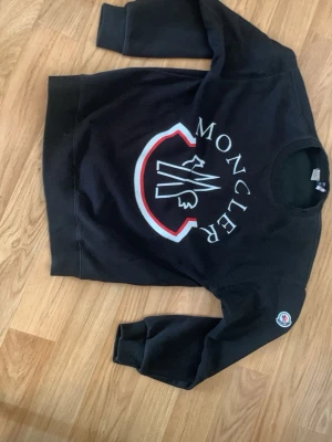 Svart Moncler sweatshirt med logga - Snygg svart sweatshirt från Moncler med stor vit och röd logga på bröstet och ett broderat Moncler-märke på ärmen. Tröjan har rund halsringning och ribbade muddar. Perfekt för en trendig och avslappnad stil.