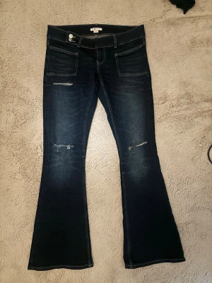 Mörkblå bootcut jeans från Gina Tricot - Snygga mörkblå bootcut jeans från Gina tricot med slitningar på låren och knäna. Jeansen har stora framfickor och klassisk femficksdesign. Materialet är stretchigt denim som sitter skönt och formar sig efter kroppen. Perfekta för en avslappnad och trendig look.