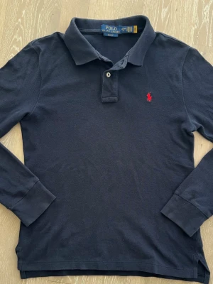 Marinblå långärmad piké Polo Ralph Lauren - Marinblå långärmad piké från Polo Ralph Lauren med klassisk krage och två knappar vid halsen. Pikén har en röd broderad logga på bröstet och är gjord i mjuk bomull. Perfekt för dig som gillar stilrena plagg med ikonisk design.
