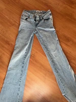 Ljusblå bootcut jeans med brodyr - Säljer ett par ljusblå bootcut jeans med snygg broderad detalj i guld på bakfickan. Jeansen har klassisk femficksmodell, låg midja och slitna tvättade detaljer. Perfekta för dig som gillar vintagevibbar och unik stil.