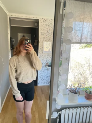 Beige cropped stickad tröja - Säljer en beige cropped stickad tröja med breda ribbstickade muddar och avslappnad passform. Tröjan har långa ärmar och är perfekt för lager på lager. Materialet känns mjukt och stretchigt, vilket gör den skön att bära.