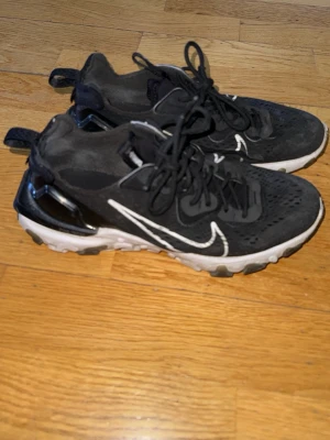 Nike React  - Svarta Nike React Element 55 sneakers med vit sula och vit swoosh på sidan. Ovandelen är i mesh och syntetmaterial med reflekterande detaljer och snörning. Sulan har ett unikt mönster och dragflik bak för enkel påtagning. Perfekta för en sportig och trendig look.