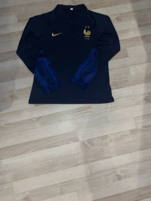 Frankrike Nike långärmad träningströja - Mörkblå långärmad träningströja från Nike med Frankrikes landslagslogga och två stjärnor på bröstet. Guldigt Nike-märke och blå mönstrade detaljer på ärmarna. Perfekt för fotbollsträning eller sportiga aktiviteter.