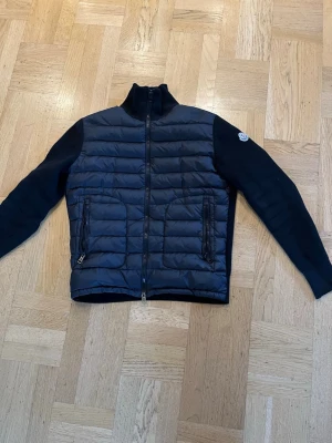 Svart cardigan från Moncler - Svart cardigan från Moncler med quiltad front och stickade ärmar. Jackan har två fickor med dragkedja och hög krage. Moncler-logga på vänster ärm. Perfekt för kalla sommardagar och snygg att bära till jeans.