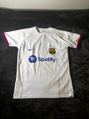 Vit FC Barcelona Nike Dri-FIT tröja - Säljer en vit FC Barcelona matchtröja från Nike med Dri-FIT material. Tröjan har klubbens emblem på bröstet, Nike-logga och Spotify som sponsor. Ärmarna har blå och röda detaljer. Perfekt för dig som älskar fotboll och vill representera Barca.