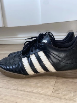 Svarta platta Adidas sneakers med vita ränder - Klassiska svarta sneakers från Adidas med de ikoniska tre vita ränderna på sidorna. Skorna har svart ovandel i läder och svart snörning, samt en brun sula. Perfekta för en sportig och stilren look. I storlek 37 1/3 
