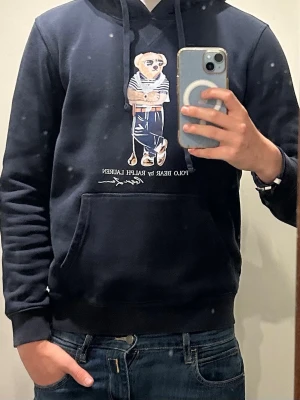 Mörkblå hoodie Polo Bear Ralph Lauren - Snygg mörkblå hoodie från Ralph Lauren med Polo Bear-tryck på bröstet. Klassisk känguruficka framtill och dragsko i huvan. Perfekt för dig som gillar streetwear och vill ha något bekvämt men ändå stilrent.