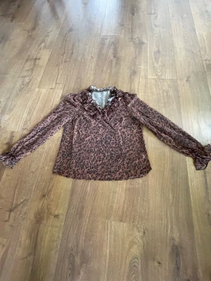 Brun leopardmönstrad blus med volang - Snygg brun blus med leopardmönster och volangdetaljer vid kragen. Blusen har volangera, långa ärmar med resår vid ärmslut och är tillverkad i ett lätt, luftig material. Perfekt för dig som gillar djurmönstrat och trendiga detaljer.💗