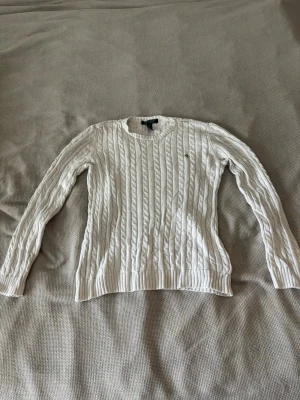 Vit kabelstickad tröja från Ralph Lauren - Säljer en vit kabelstickad tröja från Ralph Lauren med broderad RL-logga på bröstet. Tröjan har rund halsringning, långa ärmar och ribbstickade muddar. Perfekt för dig som gillar klassisk och stilren design.