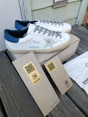 Golden Goose vita sneakers med blå detalj - Golden Goose sneakers i vitt läder med grå mockastjärna på sidan, grå skosnören och blå hälkappa. Klassisk låg modell med beige sula och GGDB/SSTAR-tryck på sidan. Snygg och unik design som sticker ut.