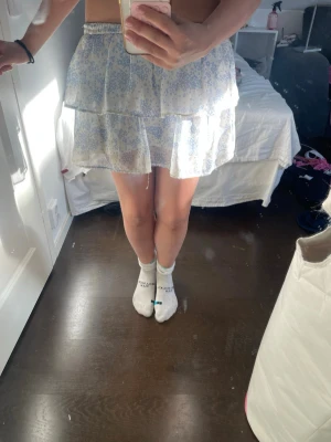 Blommig ljusblå volangkjol - Söt kort kjol med ljusblå blommönster och volangdetaljer. Kjolen har en elastisk smock i midjan och är gjord i ett lätt och luftigt material, perfekt för varma dagar. Passformen är normal och kjolen har en feminin och ungdomlig stil.