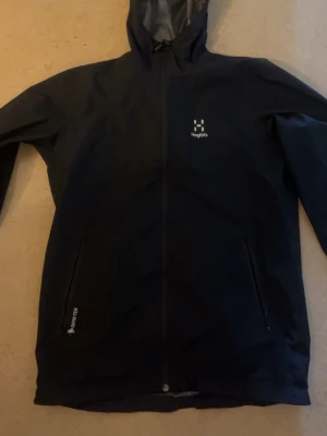 Svart vindjacka från Haglöfs Gore-Tex - Svart vindjacka från Haglöfs med hel dragkedja, huva och två sidofickor. Tillverkad i slitstarkt Gore-Tex-material som står emot vind och regn. Diskret Haglöfs-logga på bröstet och justerbar huva för extra skydd.