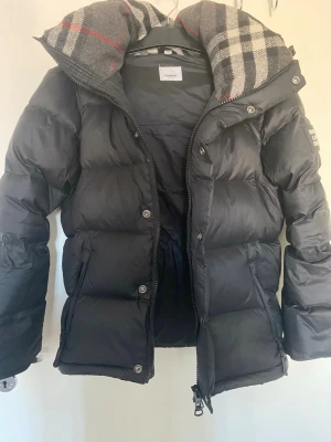 Svart pufferjacka från Burberry - Snygg svart pufferjacka från Burberry med klassiskt rutigt foder i huvan. Jackan har dragkedja och knappar framtill, två sidofickor med dragkedja och en hög krage. Perfekt för kalla vinterdagar och riktigt trendig look.