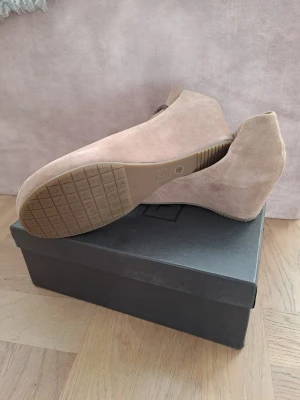 Italy Beige mocka-pumps i äkta läder  - Snygga pumps i beige mocka med kilklack och öppen tå. Skorna har ett justerbart spänne vid ankeln och en mjuk, matt finish. Perfekta för dig som vill ha både stil och komfort. 