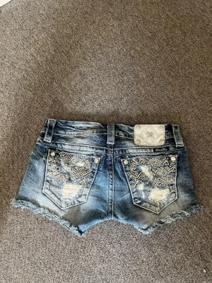 Blå jeansshorts med pärlor Miss Me - Säljer ett par blå jeansshorts från Miss Me med slitna detaljer, fransig kant och pärlbroderier på bakfickorna och sidorna. Shortsen har låg midja och klassisk femficksdesign. Perfekta för dig som vill sticka ut lite extra i sommar!