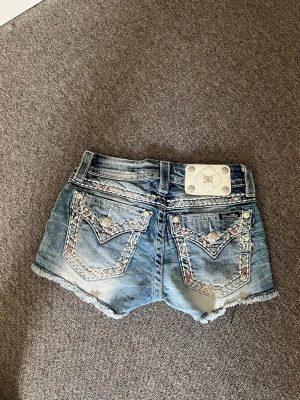 Blå jeansshorts från Miss Me - Snygga blå jeansshorts från Miss Me med slitna kanter och detaljerade bakfickor med broderier och silvriga knappar. Klassisk femficksmodell och låg midja. Perfekta för sommaren och ger en cool, avslappnad vibe.
