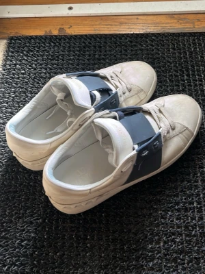 Valentino Garavani - Snygga vita sneakers från Valentino med bred mörkblå rem över snörningen. Skorna har klassisk låg profil och är tillverkade i läder med gummisula. Perfekta för dig som vill ha en stilren och exklusiv look.