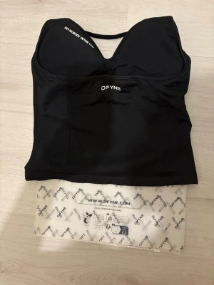 Ny top Dfyne Impact - Svart bodytop från DFYNE med smala axelband och lätt vadderad byst. Tillverkad i stretchigt material som formar sig efter kroppen. Diskret DFYNE-logga framtill och snygg rynkdetalj vid bysten. Perfekt för dig som vill ha en stilren och bekväm look.