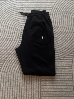 Svarta mjukisbyxor från Polo Ralph Lauren - Svarta mjukisbyxor från Polo Ralph Lauren med klassisk vit logga på benet. Byxorna har resår och dragsko i midjan samt muddar vid bensluten. Perfekta för en avslappnad stil. Tillverkade i mjukt material för extra komfort.