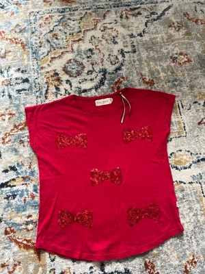 Röd topp med paljettrosetter  - Snygg röd topp från Lindex med fem glittriga paljettrosetter framtill. T-shirten har en enkel, ärmlös design och rund halsringning. Perfekt för dig som vill sticka ut med lite extra glitter. 