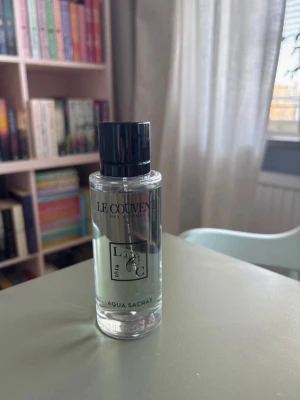 Le Couvent Aqua Sacrae parfym - Fräsch parfym från Le Couvent med namnet Aqua Sacrae. Flaskan är genomskinlig med svart plastkork och stilren etikett. Doften är elegant och flaskan har en modern, minimalistisk design som passar perfekt på sminkbordet.