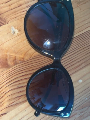 Svarta solglasögon från Michael Kors - Snygga svarta solglasögon från Michael Kors med stora, runda bågar och mörka glas. Klassisk design med diskret guldfärgad MK-logga på skalmarna. Perfekta för att lyfta din stil under soliga dagar.