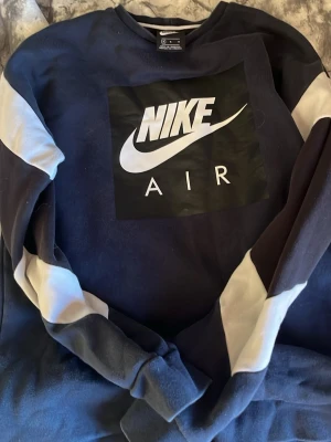 Nike Air blå sweatshirt med tryck - Snygg blå sweatshirt från Nike Air med stort vitt logotryck på bröstet. Tröjan har kontrasterande ljusare partier på ärmarna och är tillverkad i mjukt bomullsmaterial. Perfekt för en sportig och avslappnad stil.